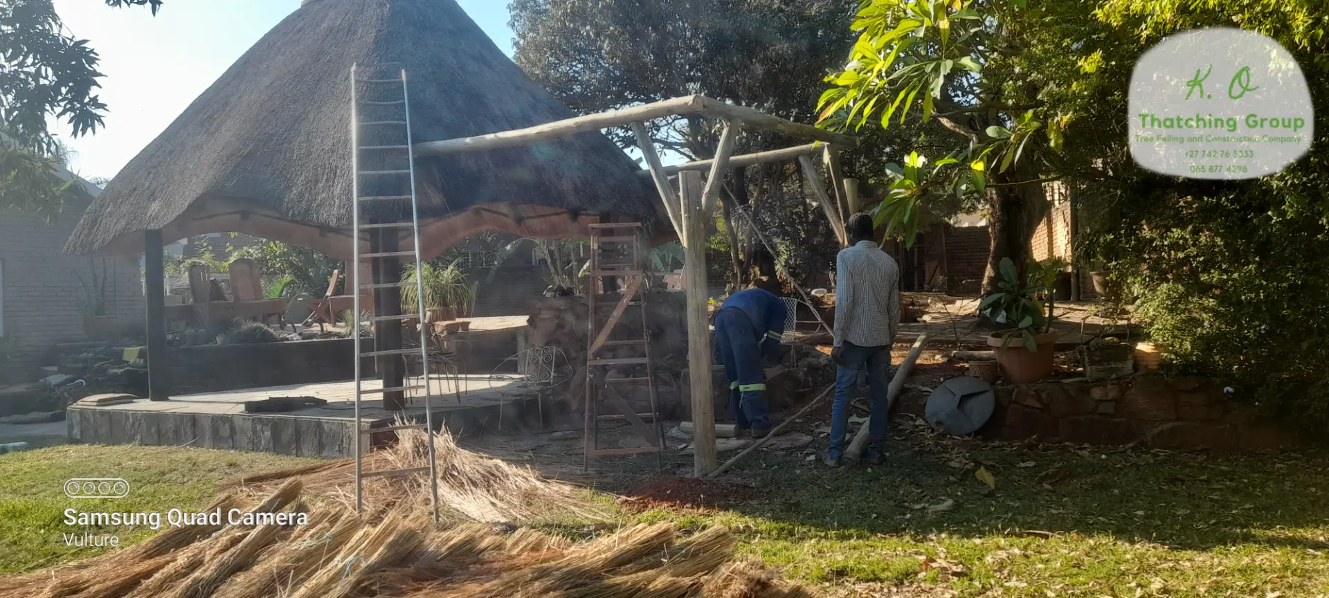 Timber pole framing and thatch bundles on a structure under construction, with crew on ladders